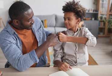 Portrait of African-American father fist bumping smiling son while doing homework together at home