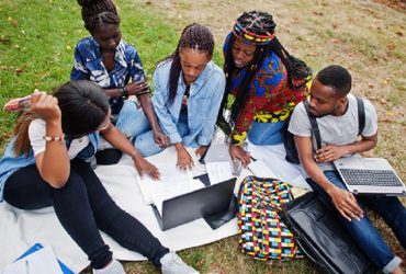 Group of five african college students spending time together on campus at university yard. Black afro friends sitting on grass and studying with laptops.